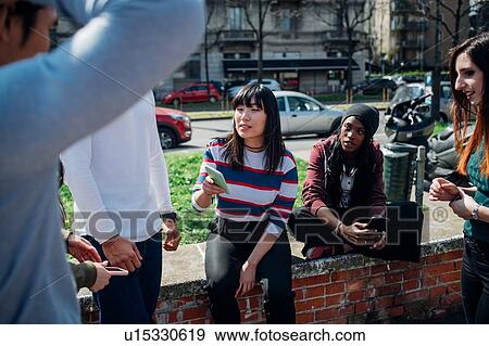 Young female and male adult friends sitting on wall chatting and looking at smartphones View Large Photo Image Stock Photo - Young female and male adult friends sitting on wall chatting and looking at smartphones. Fotosearch