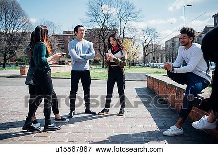 Stock Photo - Young female and male adult friends sitting on wall chatting and looking at smartphones. Fotosearch