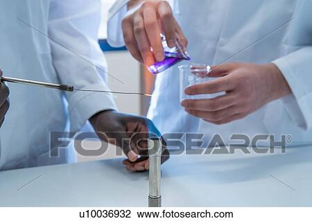 Stock Image - Young female and male scientists experimenting with liquid and bunsen burner in laboratory, cropped. Fotosearch
