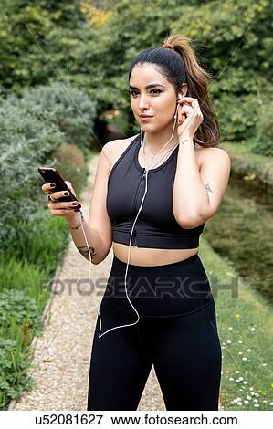 Stock Photo - Young female runner inserting earphones on riverside path. Fotosearch