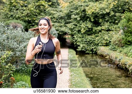 Stock Image - Young female runner listening to earphones while running on riverside path. Fotosearch