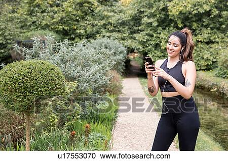 Stock Image - Young female runner selecting earphone music on riverside path. Fotosearch