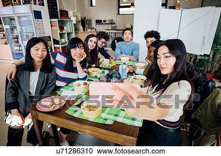 Stock Image - Young male and female business team having working lunch at cafe table, taking smartphone selfie. Fotosearch