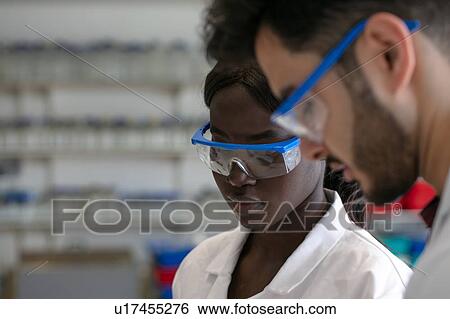Stock Photograph - Young male and female scientists wearing protective goggles in laboratory, close up. Fotosearch