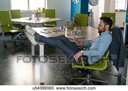 Young male business creative with feet up looking at smartphone in office View Large Photo Image Stock Image - Young male business creative with feet up looking at smartphone in office. Fotosearch