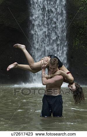 Stock Image - Young man carrying young woman over his shoulder in river by Tibumana Waterfall in Bali, Indonesia. Fotosearch