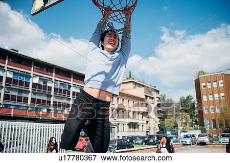 Stock Photo - Young man hanging from basketball hoop at city basketball court. Fotosearch