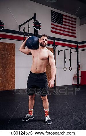 Stock Image - Young man training, carrying atlas ball on shoulder in gym. Fotosearch