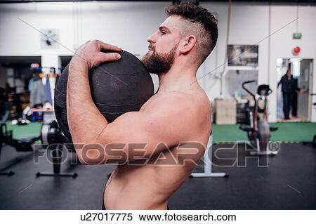 Stock Photography - Young man training, lifting atlas ball in gym, side view. Fotosearch