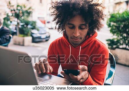 Stock Photography - Young man using smartphone and laptop at cafe. Fotosearch
