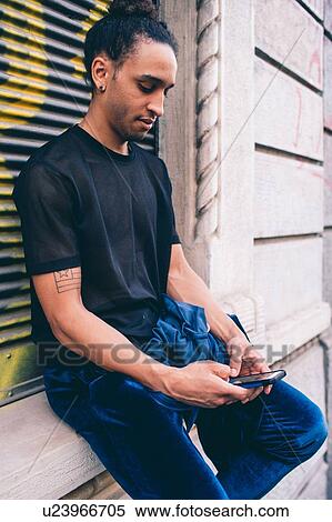 Stock Photography - Young man using smartphone on window ledge of building. Fotosearch
