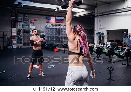 Stock Image - Young woman and man training, lifting dumbbells in gym. Fotosearch