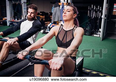 Stock Image - Young woman and man training on rowing machines together in gym. Fotosearch