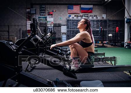 Stock Photograph - Young woman and man training on rowing machines together in gym, side view. Fotosearch