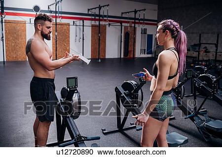Stock Image - Young woman and man training together, preparing to use rowing machines in gym. Fotosearch