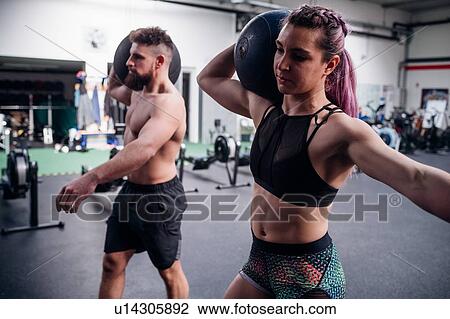 Stock Image - Young woman and man training together, carrying atlas ball on shoulders in gym. Fotosearch