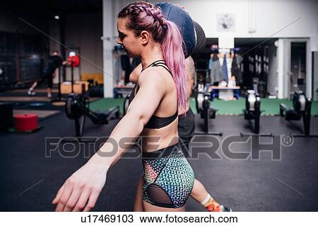 Stock Image - Young woman and man training together, carrying atlas ball on shoulders in gym, side view. Fotosearch