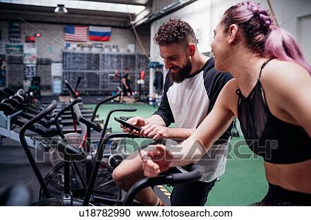 Stock Image - Young woman and man training together on gym exercise bikes, looking at smartphone. Fotosearch