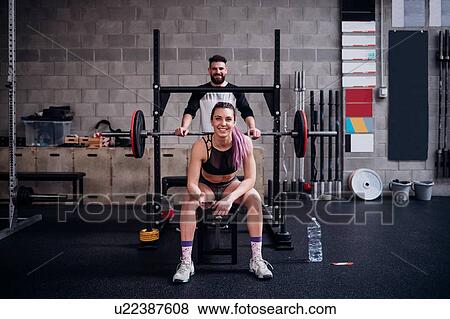 Stock Photo - Young woman and man training together in gym, sitting on weight bench, portrait. Fotosearch