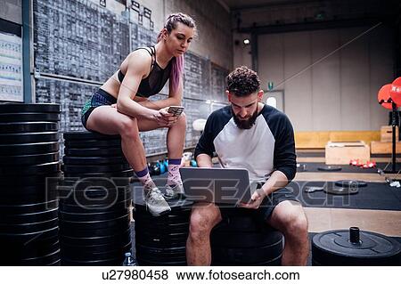 Stock Photo - Young woman and man training together in gym, sitting on weights looking at laptop. Fotosearch