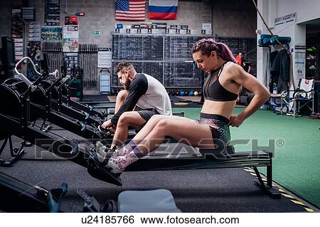Stock Photograph - Young woman and man training together, preparing to use rowing machines in gym. Fotosearch