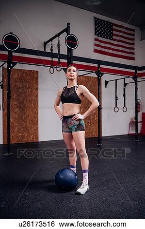 Stock Photograph - Young woman atlas ball training, standing in gym with hands on hips, full length. Fotosearch