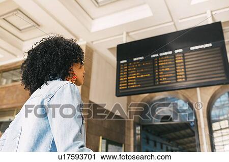 Stock Image - Young woman by arrival board in railway station. Fotosearch