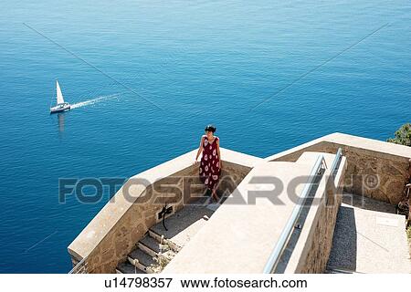 Stock Photo - Young woman enjoying sunny day by sea, sailboat in background, Alghero, Sardegna, Italy. Fotosearch