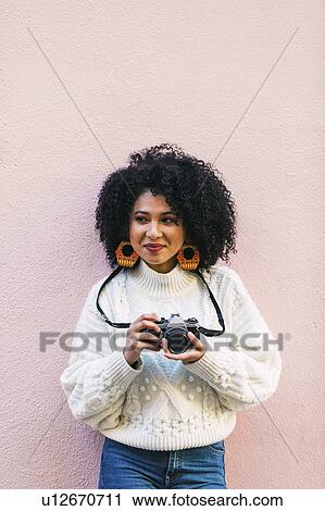 Stock Image - Young woman holding camera against pink wall. Fotosearch