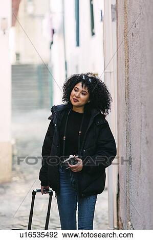 Stock Image - Young woman holding camera and suitcase in alley. Fotosearch