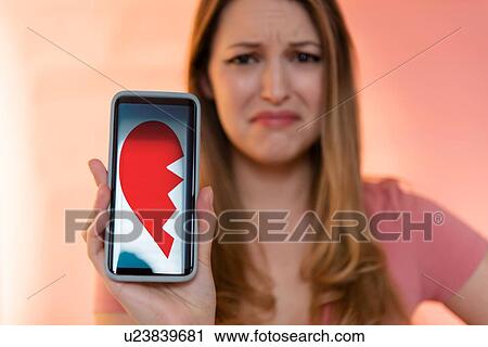 Stock Image - Young woman holding smart phone displaying broken heart. Fotosearch