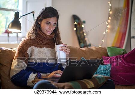 Picture - Young woman in knit sweater relaxing on living room sofa with drink and using laptop. Fotosearch