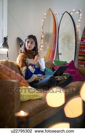 Stock Image - Young woman in knit sweater relaxing on living room sofa with drink. Fotosearch