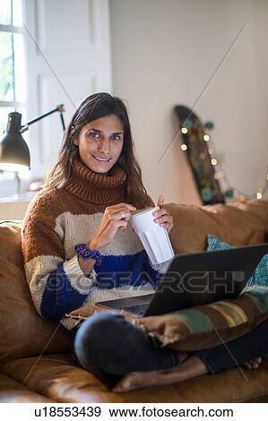 Stock Photo - Young woman in knit sweater relaxing on living room sofa with laptop and drink, portrait. Fotosearch