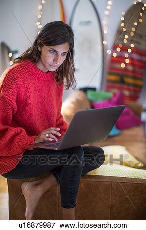 Stock Photo - Young woman in red sweater on living room sofa looking at laptop. Fotosearch