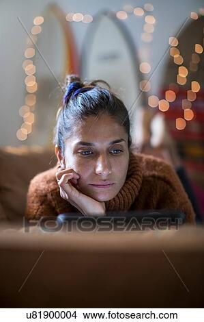 Picture - Young woman lying on living room sofa gazing at laptop, shallow focus. Fotosearch
