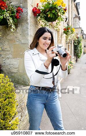 Stock Photo - Young woman on village street reviewing photos on digital camera, Cotswolds, England. Fotosearch
