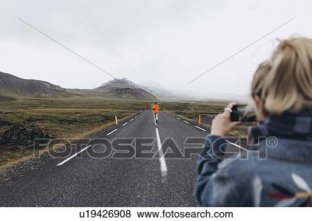 Stock Photo - Young woman photographing boyfriend walking on highway in Iceland. Fotosearch