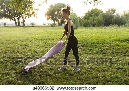 Stock Image - Young woman placing yoga mat in park. Fotosearch