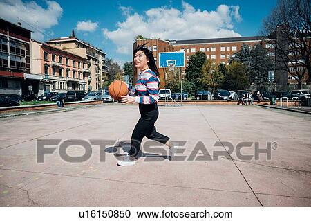 Stock Image - Young woman playing basketball on city basketball court. Fotosearch