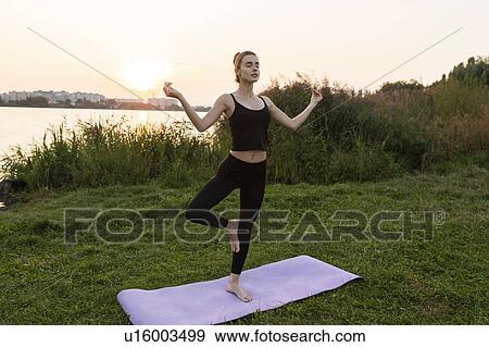 Stock Photo - Young woman practicing yoga in park at sunset. Fotosearch