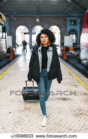 Stock Image - Young woman pulling suitcase in railway station. Fotosearch