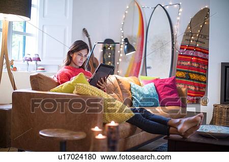Stock Image - Young woman relaxing on living room sofa looking at digital tablet. Fotosearch