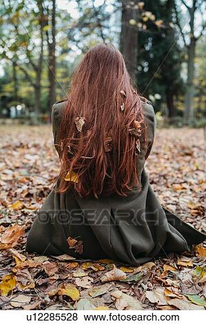 Stock Photo - Young woman sitting on leaves in park with autumn leaves tangled in long red hair, rear view. Fotosearch