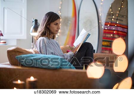 Stock Photo - Young woman sitting on living room sofa reading a book. Fotosearch