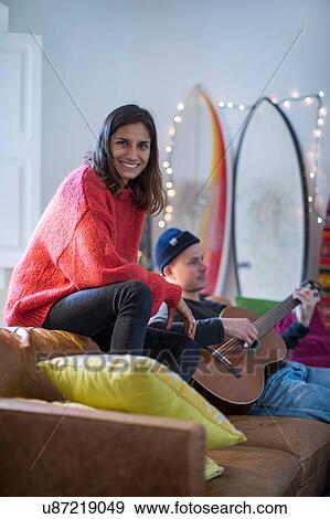 Stock Photo - Young woman sitting up on living room sofa with young man playing guitar. Fotosearch