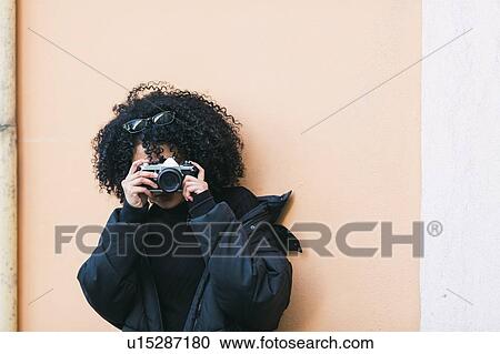 Stock Image - Young woman taking photograph against orange wall. Fotosearch