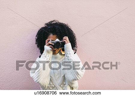 Stock Photograph - Young woman taking photograph against pink wall. Fotosearch