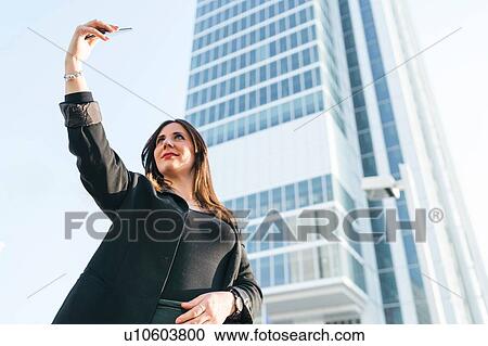 Stock Image - Young woman taking smartphone selfie in front of skyscraper, low angle view, Turin, Piemonte, Italy. Fotosearch