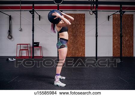 Picture - Young woman training, carrying atlas ball on shoulder in gym. Fotosearch